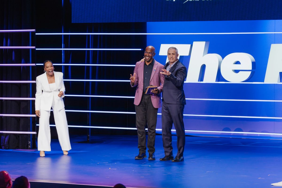 Three people on blue-lit stage, two men embracing while woman in white suit applauds at conference venue