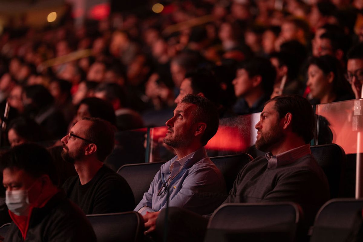 Attendees seated in red lighting watch a presentation at a conference, with a large crowd visible in.