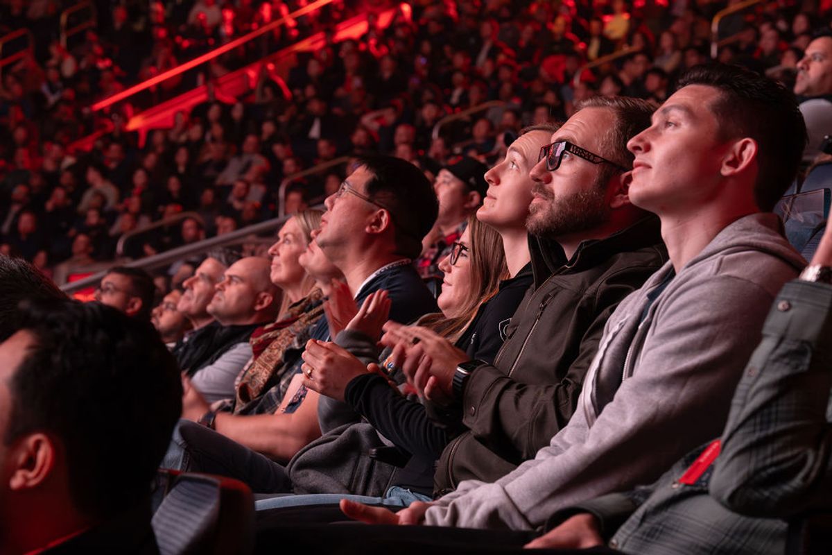 Conference attendees sitting in tiered seating, looking up attentively during a presentation with red stage.