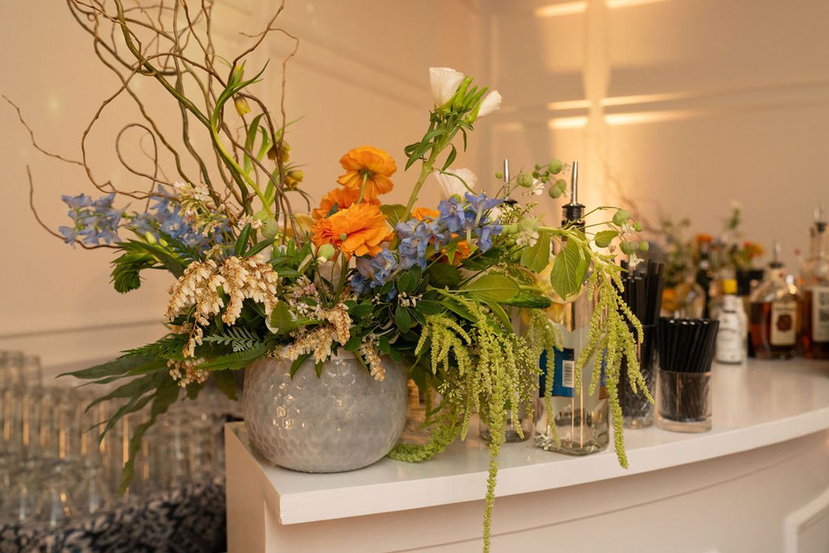 Floral arrangement with orange and blue flowers in gray vase on white shelf next to bottles at reception venue