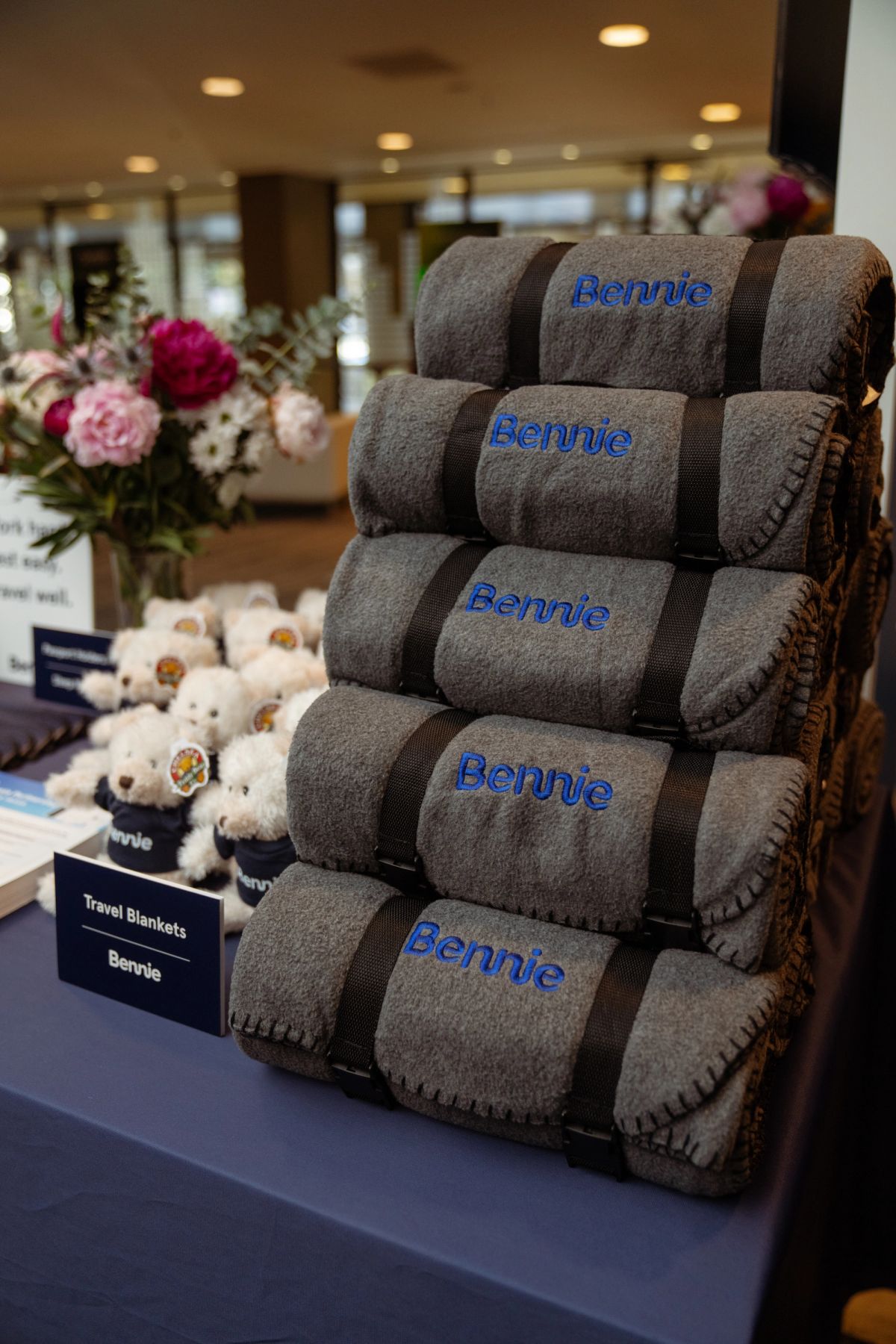 Stacked Bennie-branded travel blankets and teddy bears displayed on a table at a conference.