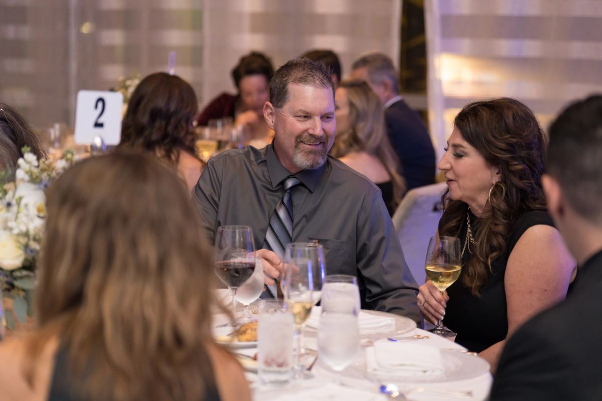 Guests in formal attire socializing at dinner tables with wine glasses and floral centerpieces at an indoor.