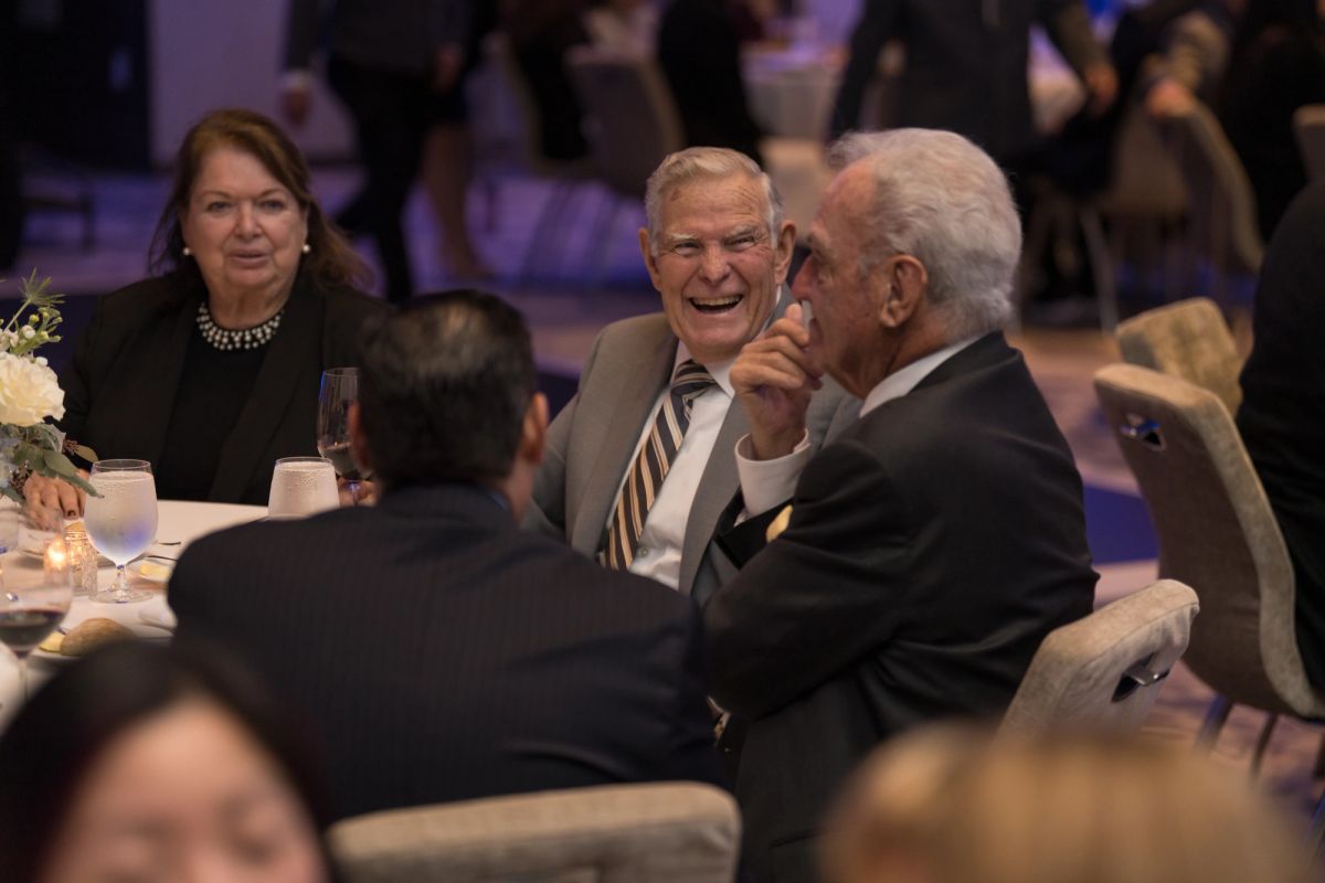 Attendees in formal attire sitting at dinner table, smiling and conversing at gala event