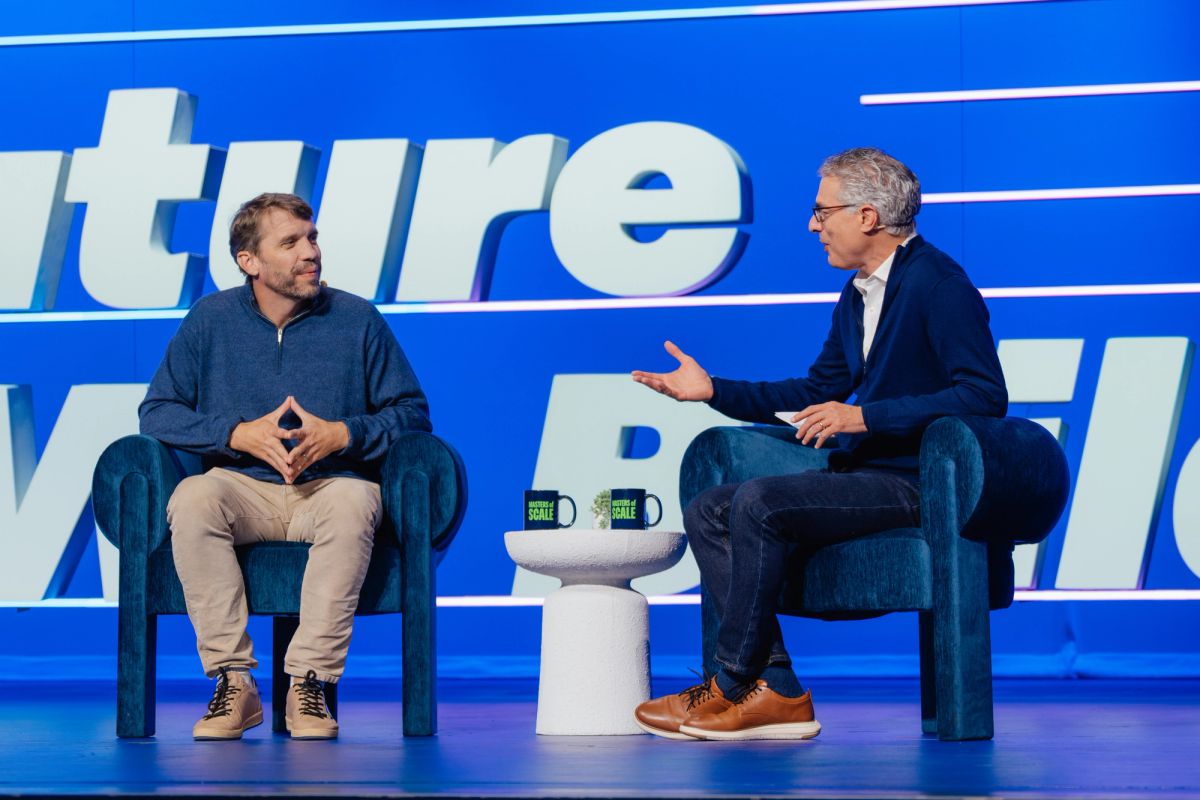 Two speakers in conversation on stage at conference, seated in blue chairs with blue backdrop displaying.
