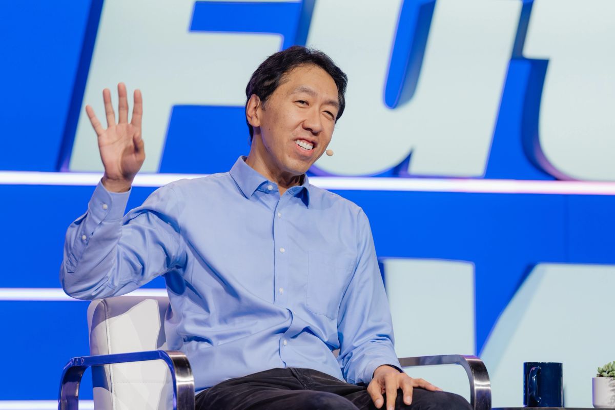 Speaker in blue shirt waving to audience while seated on stage at conference with blue backdrop