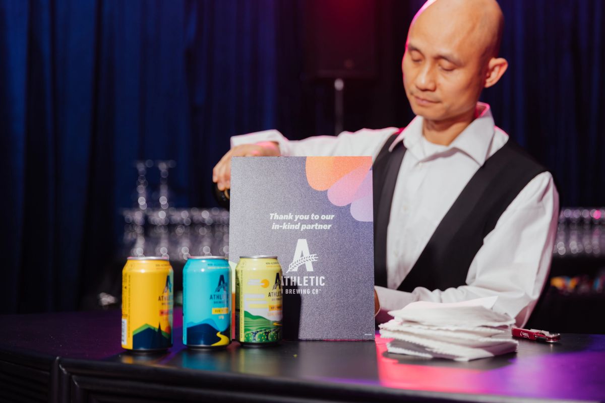 Speaker at podium holding Athletic Brewing Co. promotional material with three colorful beer cans displayed