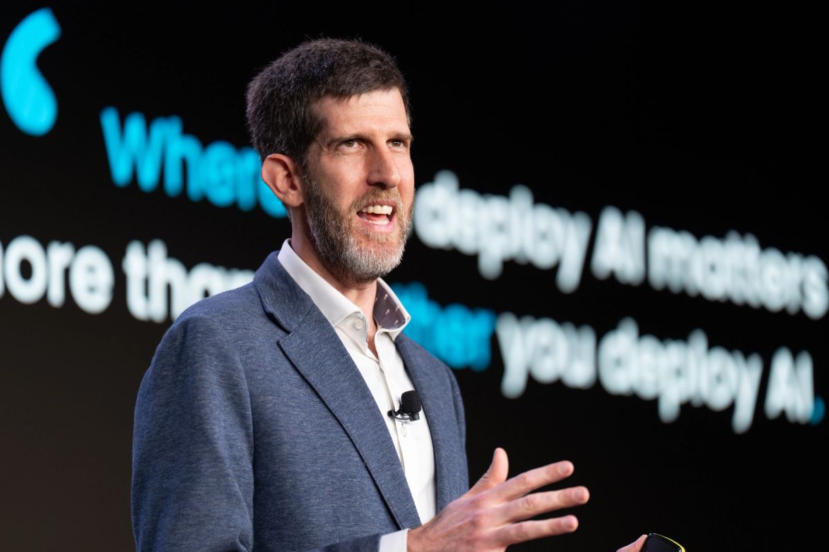 Speaker presenting on stage with blue and white text displayed on screen behind him