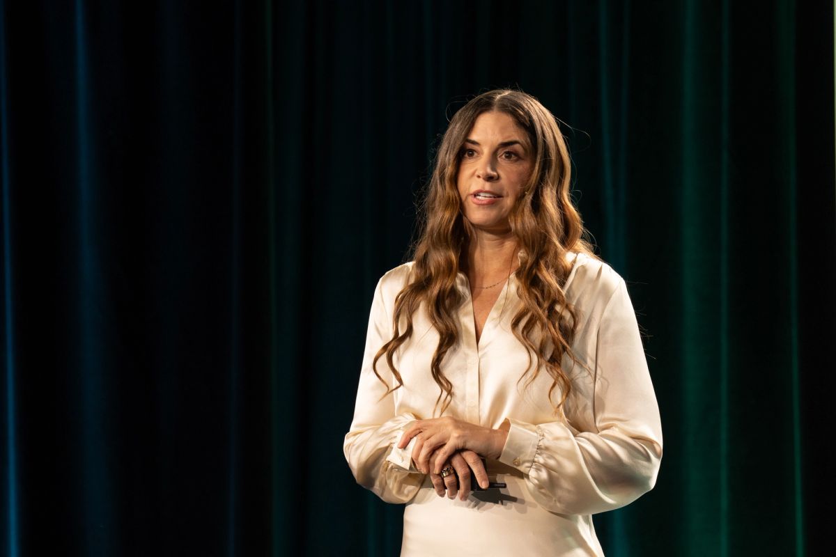 Woman in cream blouse speaking on stage before teal curtain backdrop.