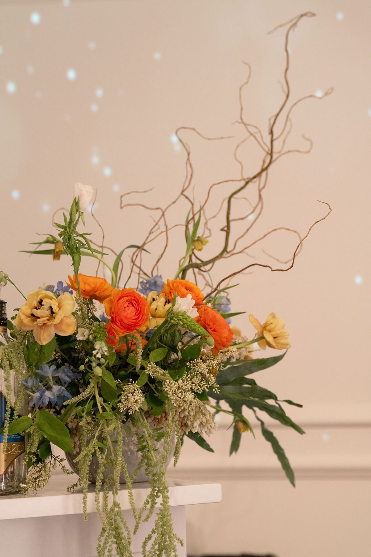 Colorful floral arrangement with orange ranunculus, yellow flowers, and curly branches on white table against.