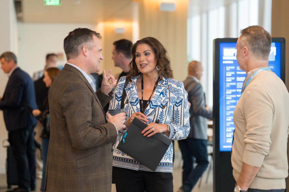 Three professionals networking in a modern conference venue lobby with digital displays in background.