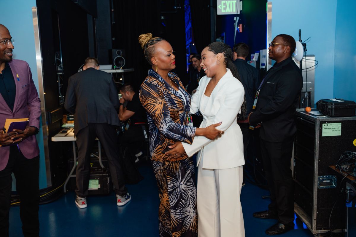 Two women networking and chatting in a blue-lit backstage area with audio equipment and other attendees