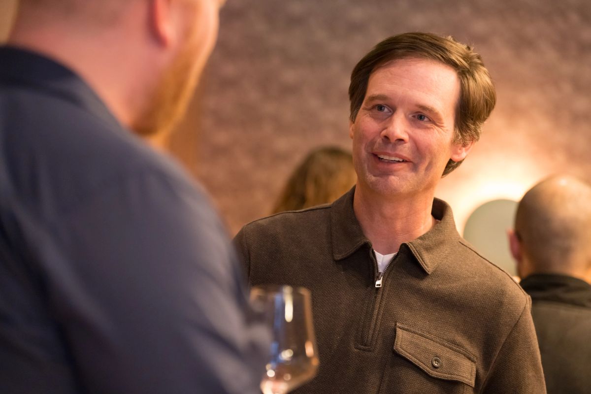 Man in brown collared shirt speaking with another person at an indoor event