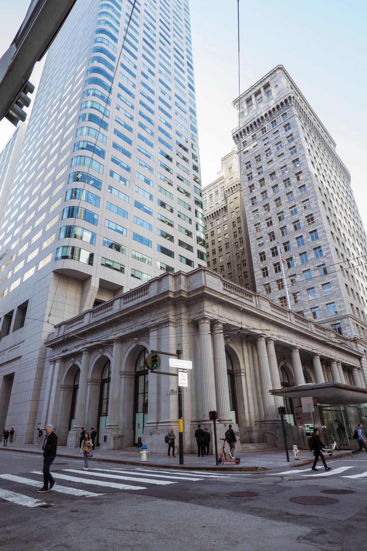 Neoclassical building at a city intersection with pedestrians, flanked by modern skyscrapers.
