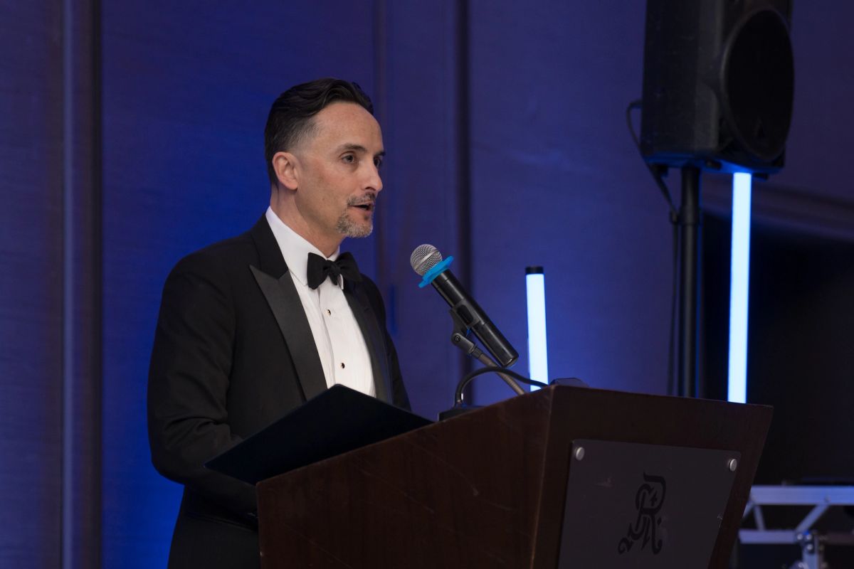 Man in black tuxedo speaking at wooden podium with microphone at formal evening event with blue lighting