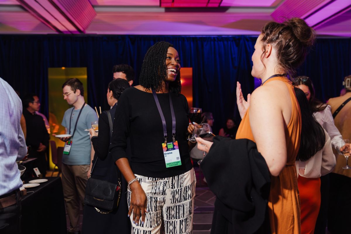 Two women networking and conversing at a conference event with colorful lighting and other attendees in.