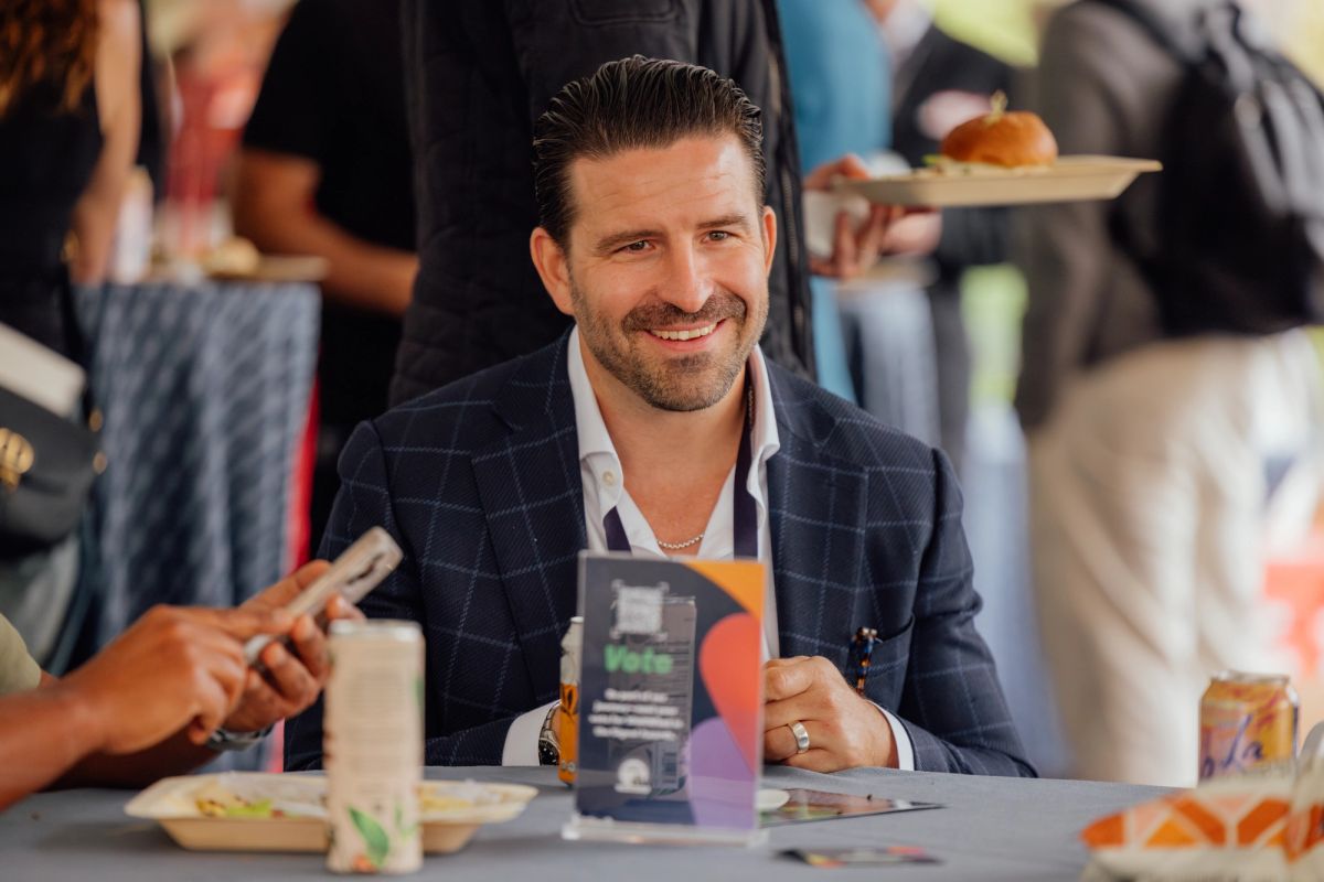 Man in navy suit jacket smiling at table during event, with attendees and food service in background.