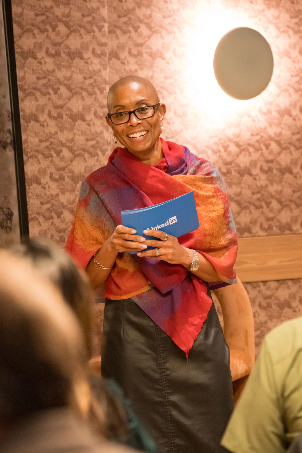 Smiling speaker in colorful shawl holds LinkedIn notebook at panel discussion, audience members visible below