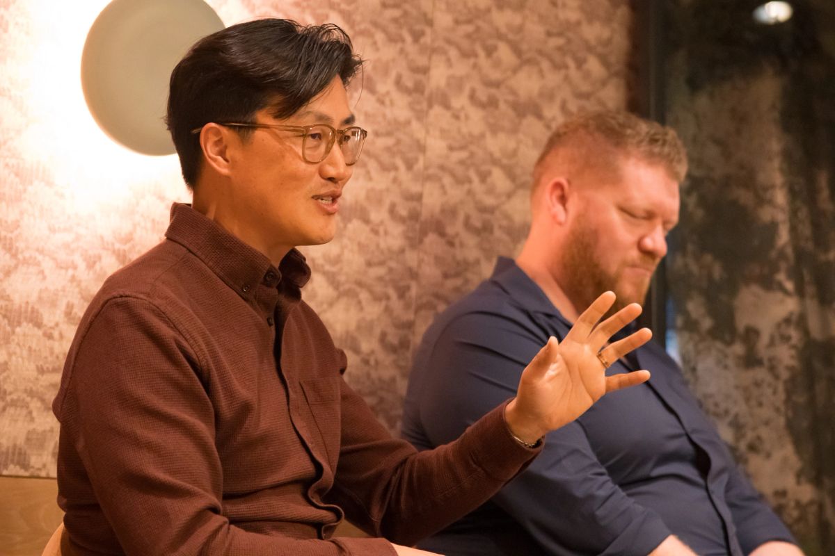 Two panelists during discussion, one speaker gesturing while talking, seated against textured wall backdrop