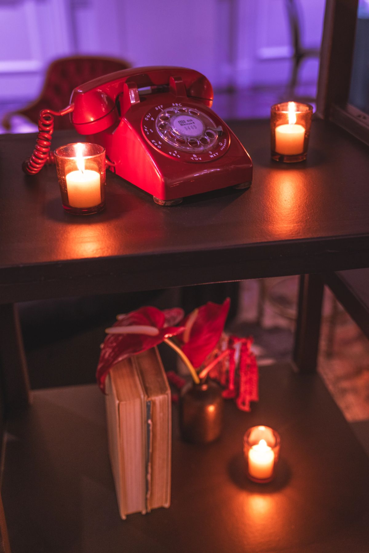 Red rotary phone on wooden table with lit candles and vintage books in moody purple lighting