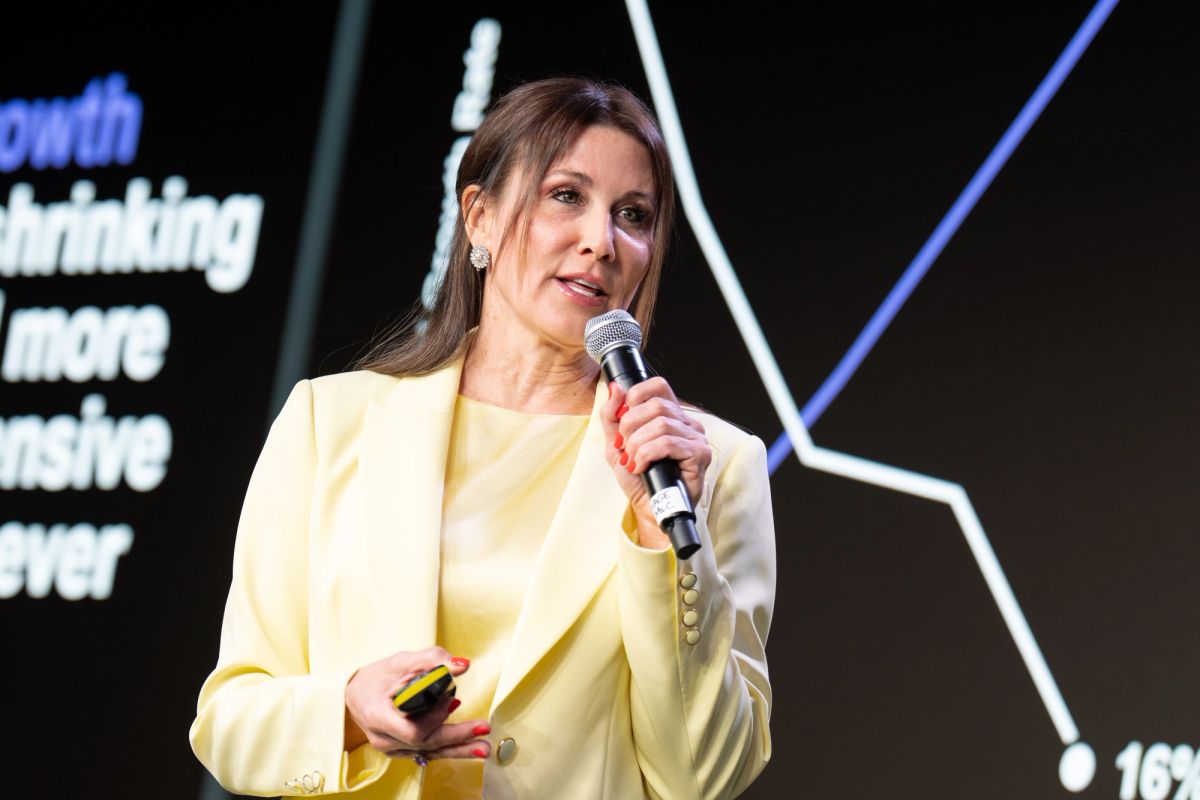 Speaker in yellow blazer presents with microphone at business conference, stage backdrop with geometric.