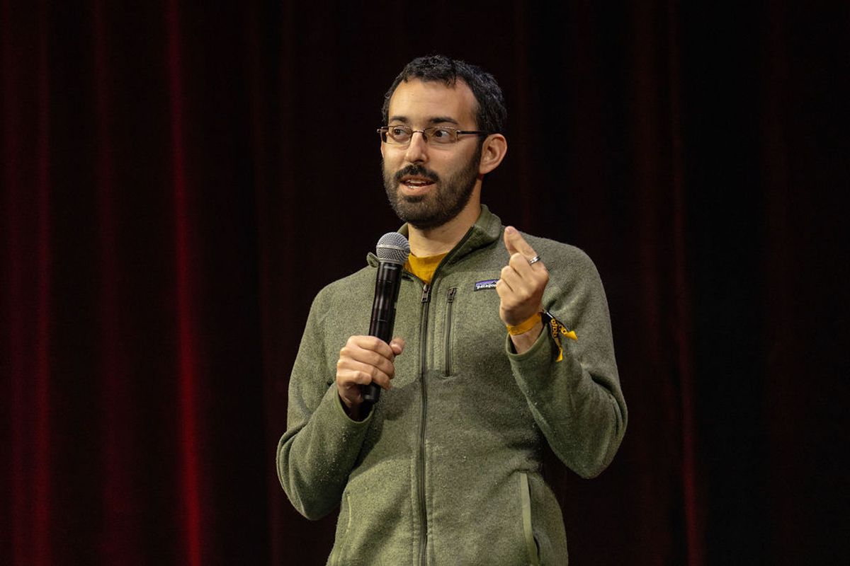 Speaker with microphone gesturing to audience on stage with dark curtain backdrop