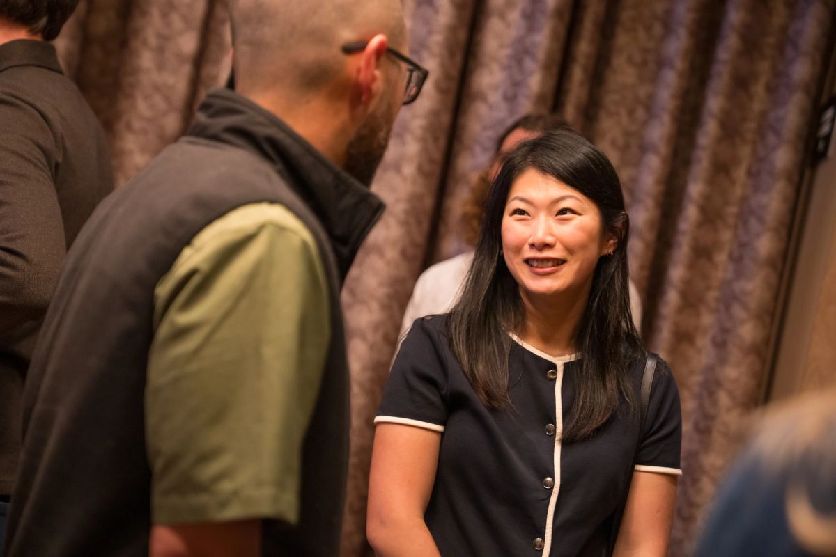 Two people conversing at an indoor event, with a woman in a dark button-up shirt smiling at a man in glasses.