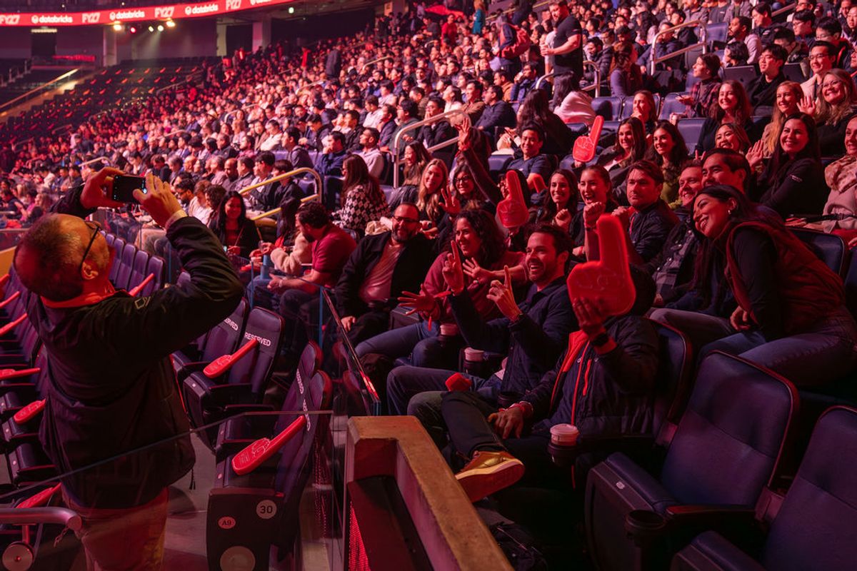 Large audience seated in arena-style seating, bathed in red and purple stage lighting, watching an event
