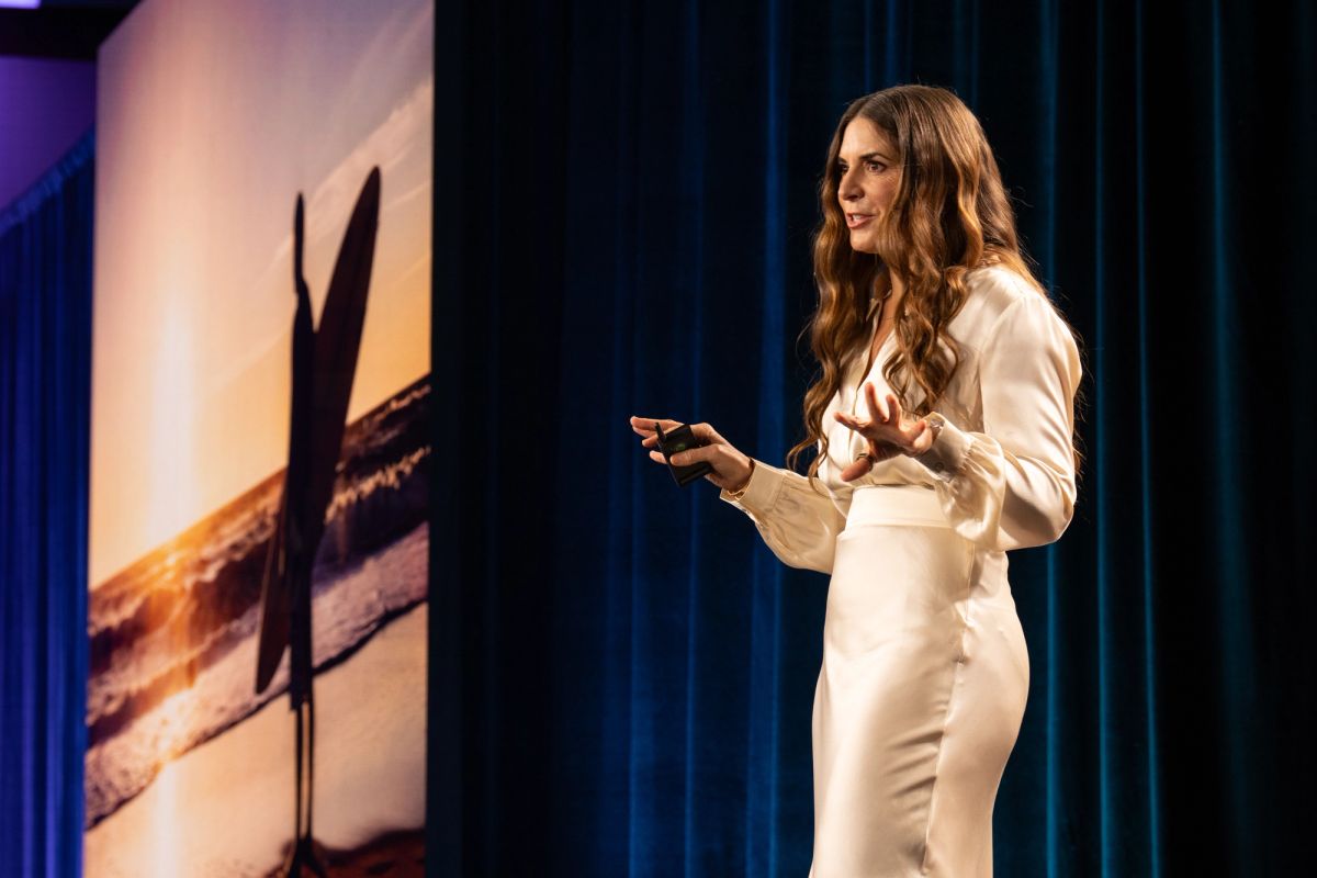 Woman in white suit presenting on stage before blue curtain backdrop.