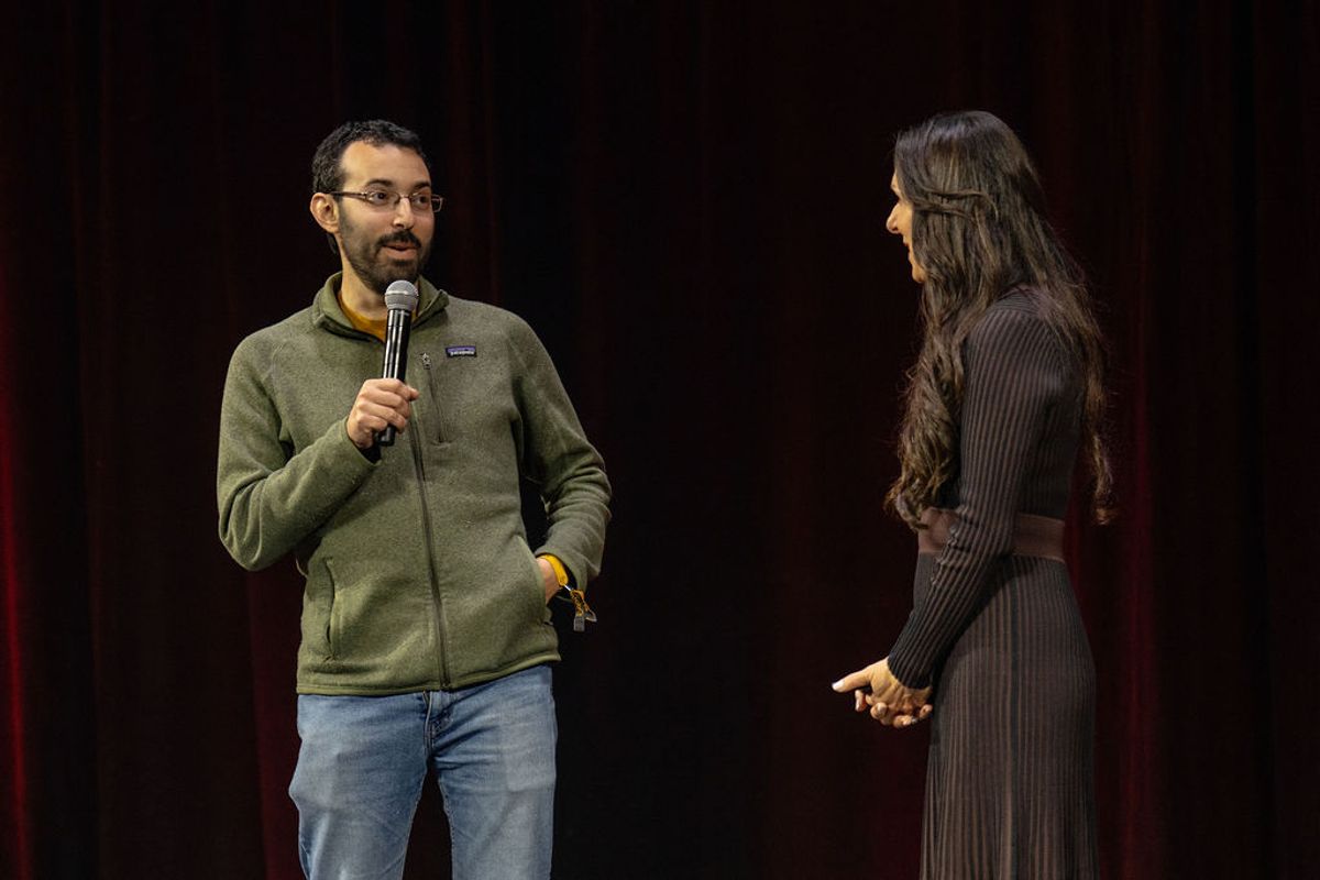 Two speakers on stage, one man holding microphone wearing green sweater, one woman in black dress