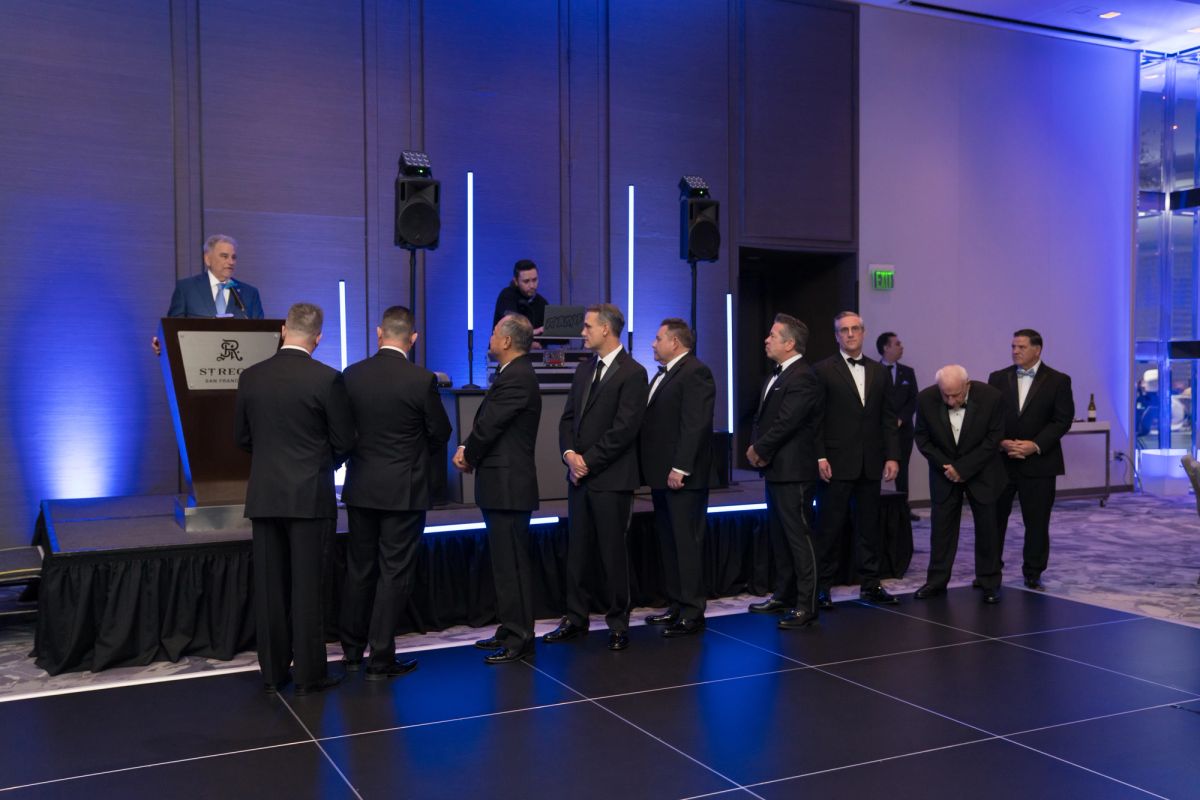 Speaker at podium addresses group of men in formal attire lined up on stage at evening gala event.