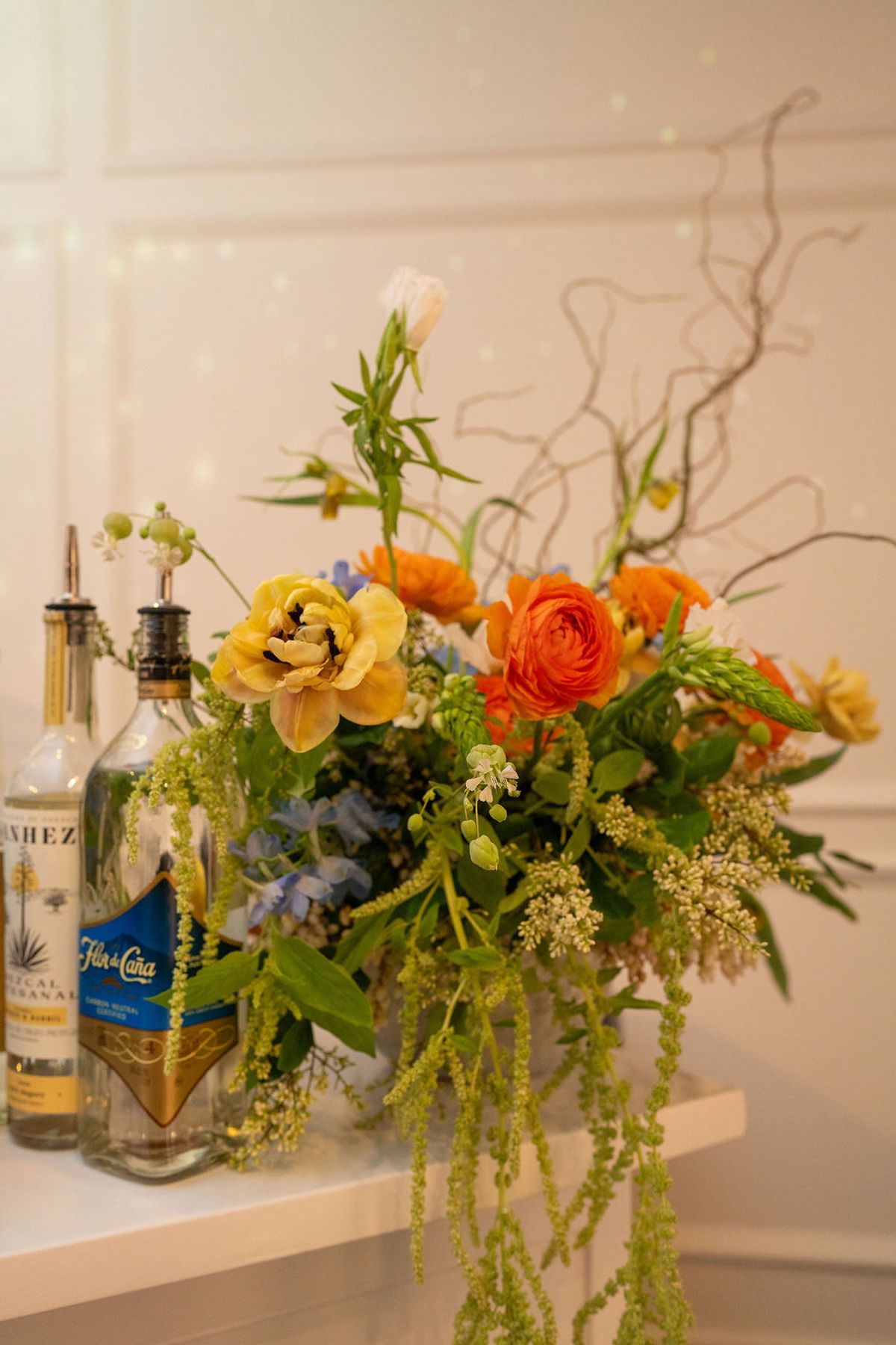 Colorful flower arrangement with yellow and orange blooms next to liquor bottles on white shelf