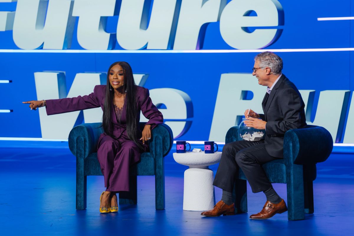 Two speakers in conversation on stage, woman in purple suit gesturing while seated in blue chairs