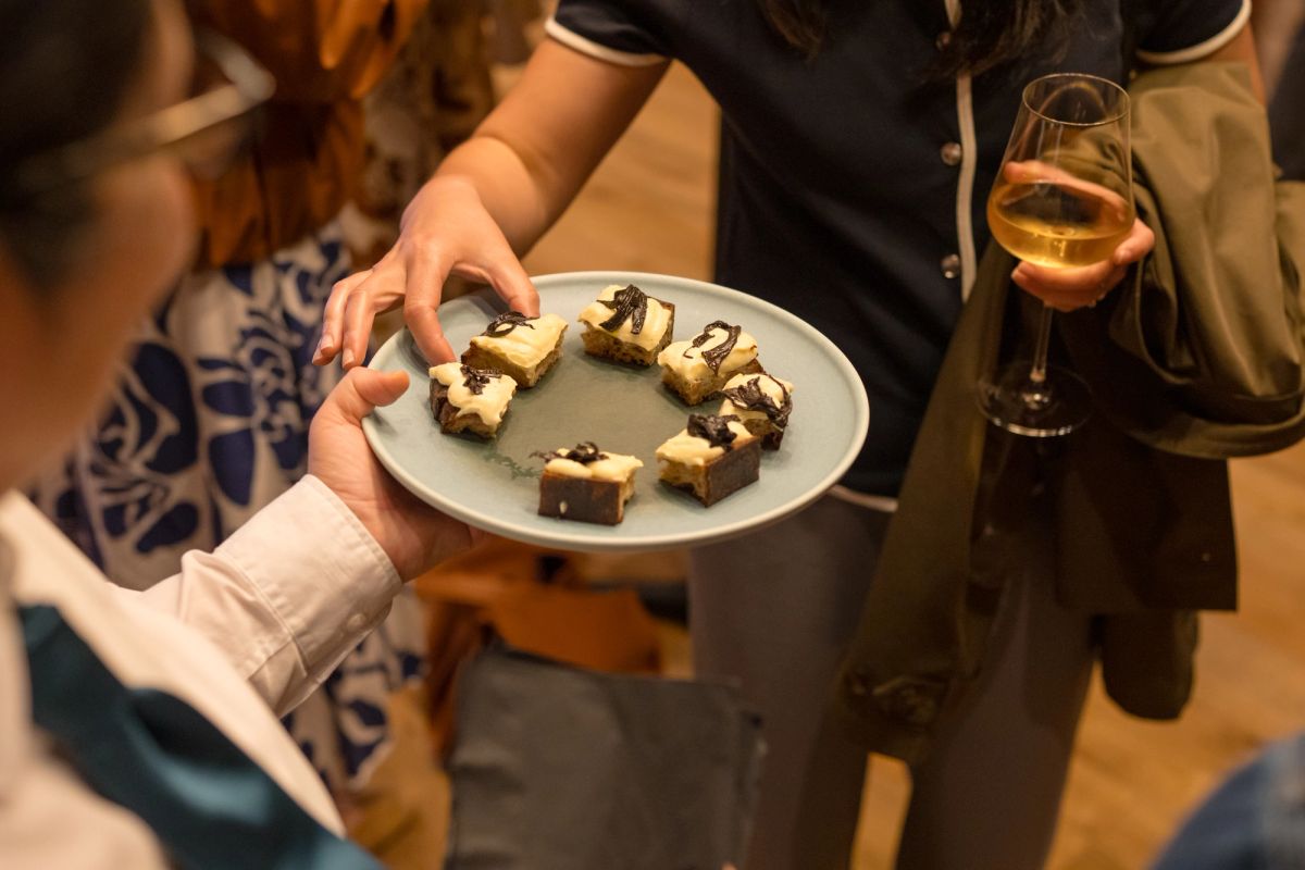 Person holding plate of small desserts while attendees hold champagne glasses at indoor event