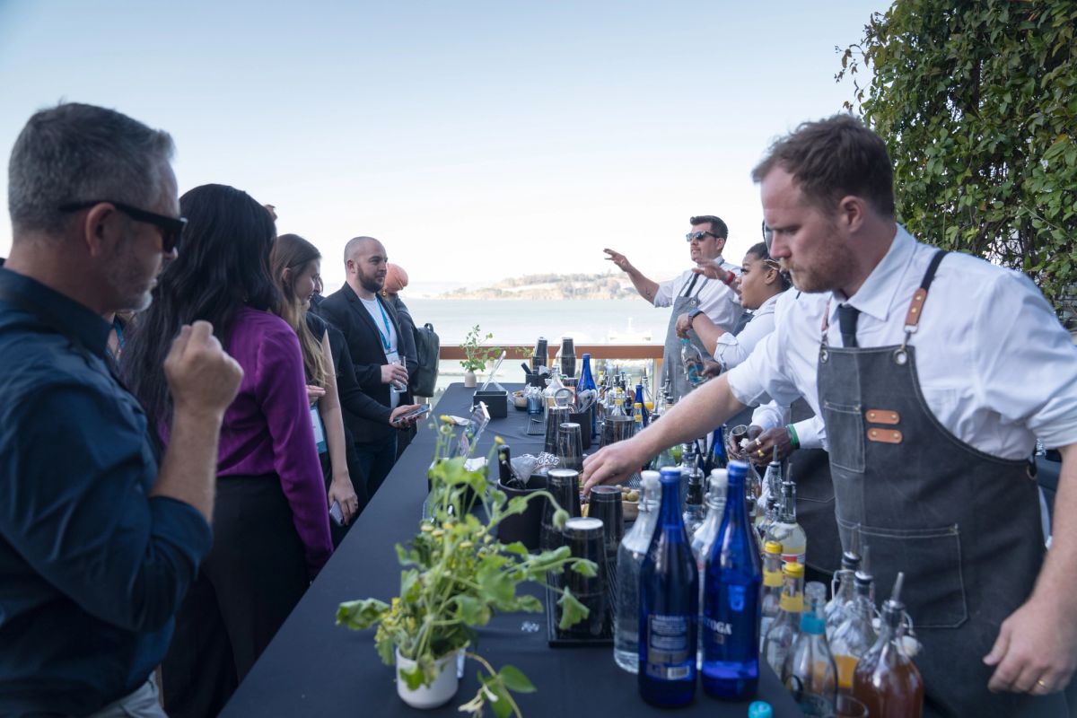 Bartender in apron serves drinks to attendees at outdoor rooftop event with city view in background.