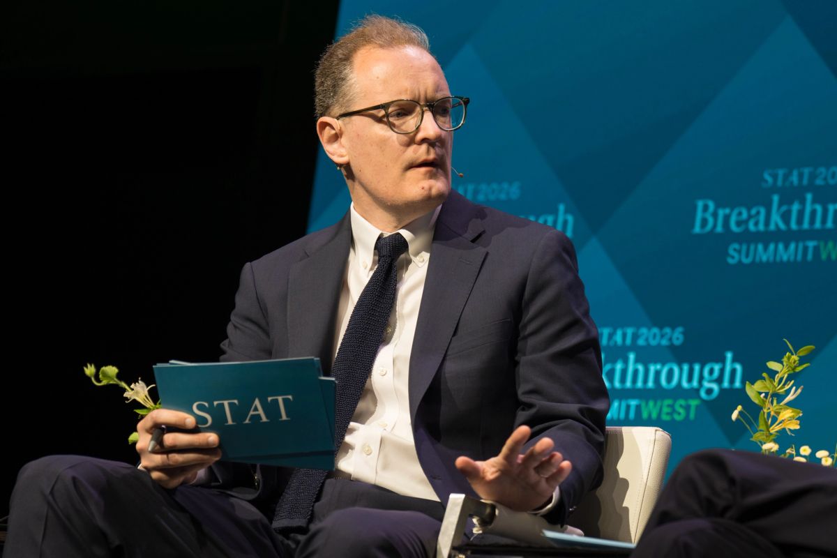 Speaker in dark suit and glasses gestures while holding materials at conference panel with blue backdrop