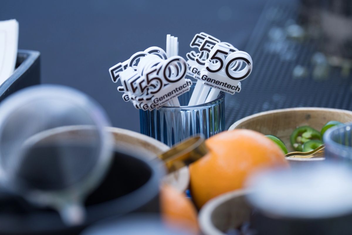 Table setting with coffee cups, fruit, and small white flags displaying "50" and "Genentech" logos