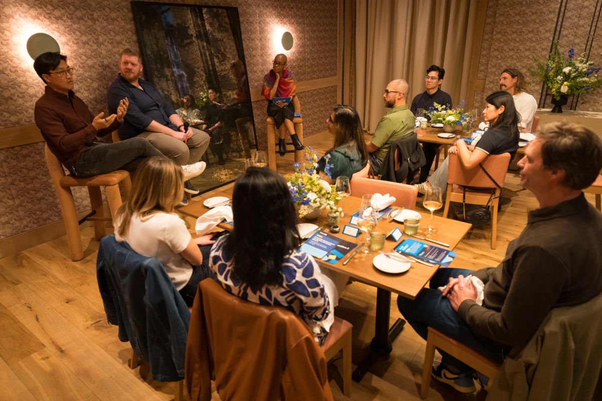 Panel discussion with speaker addressing seated audience in modern room with wooden tables and warm lighting