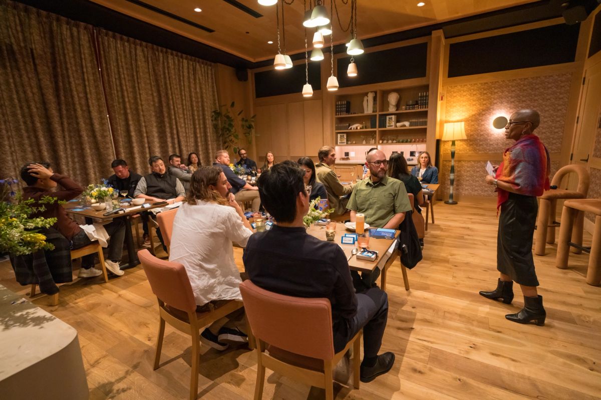 Speaker addresses seated audience around wooden table in warm, modern conference room with pendant lighting.