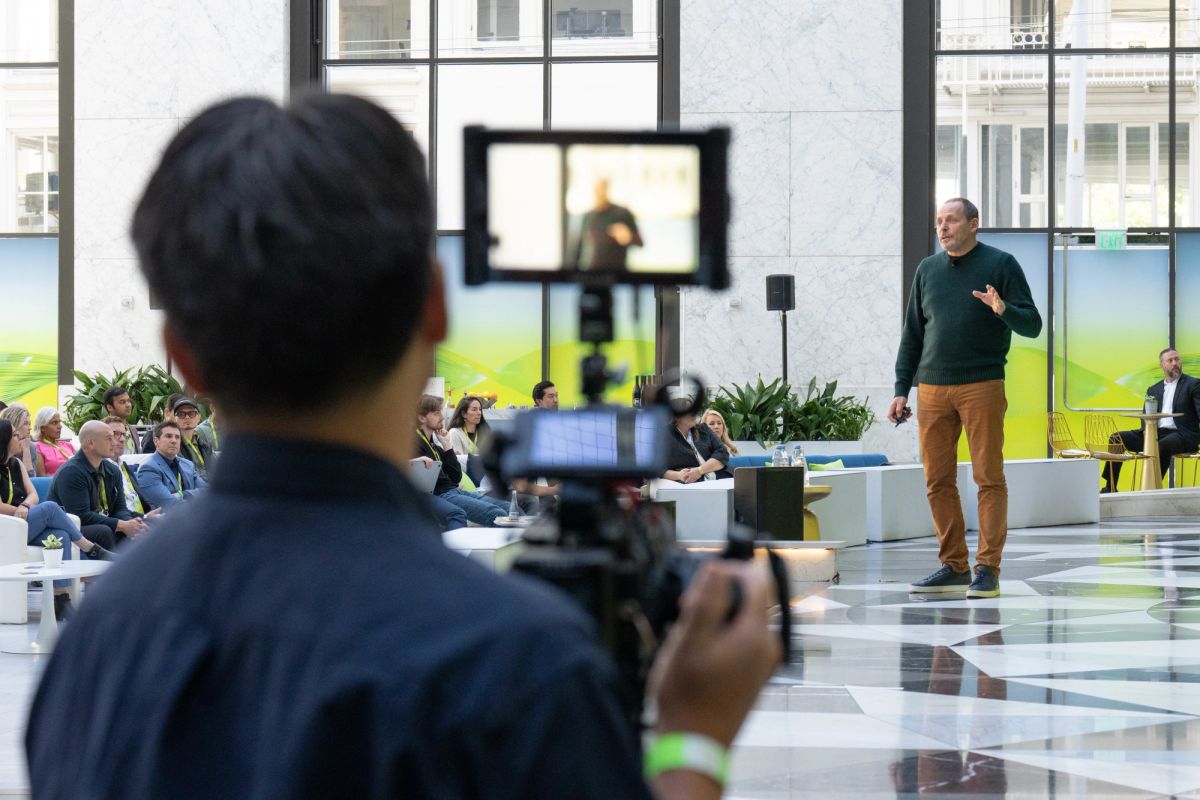 Videographer films a speaker presenting to seated attendees in a modern atrium conference space.