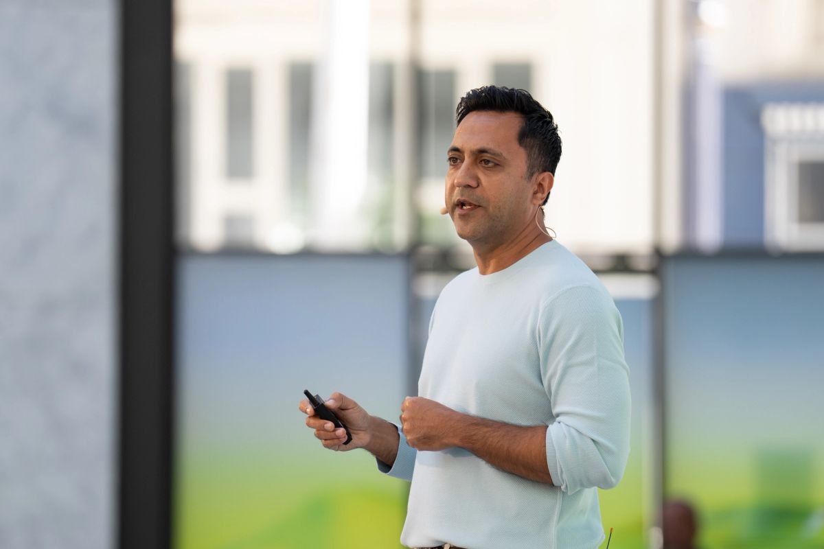 Speaker presenting outdoors, holding a phone, wearing a light sweater, with blurred building backdrop.