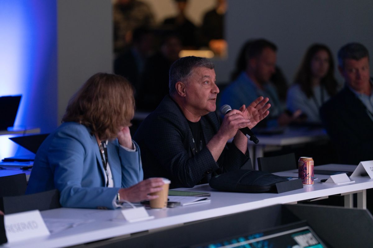 Two panelists at conference table, one speaking into microphone while gesturing, audience members visible in.