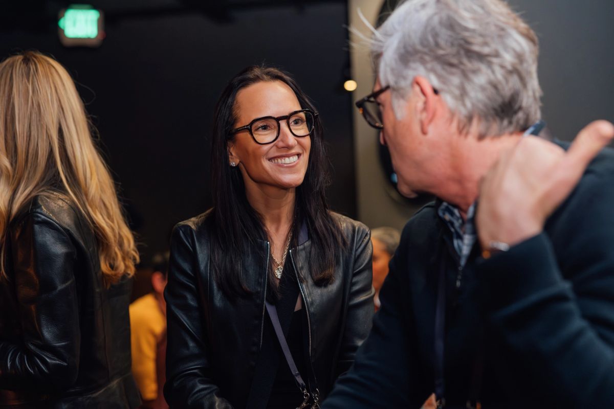 Two people in conversation at an indoor event, woman in black leather jacket and glasses smiling at man