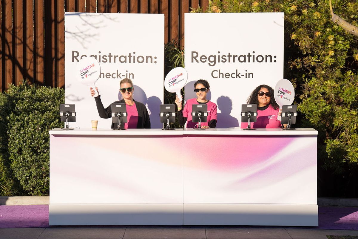 Three women in pink sit at registration check-in table with white backdrop, holding circular signs outdoors