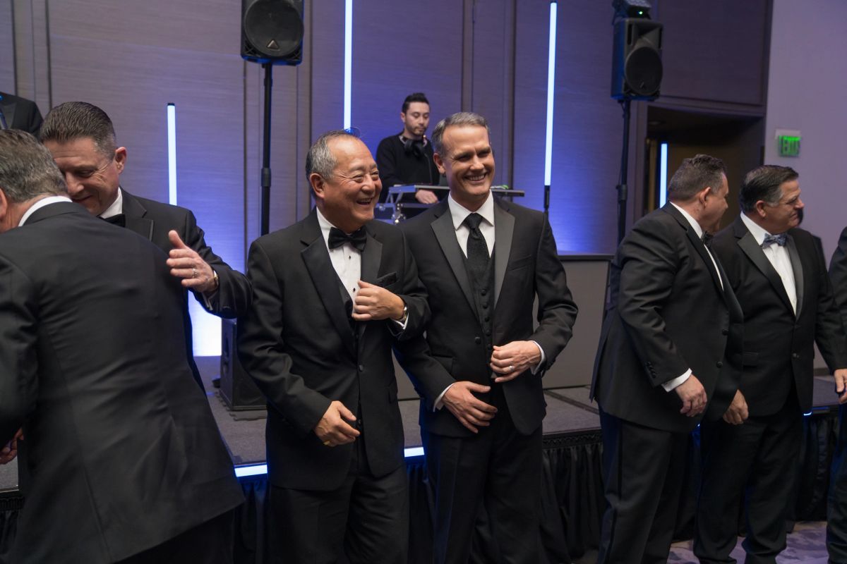 Men in tuxedos and suits socializing at a formal evening event with purple lighting and speakers