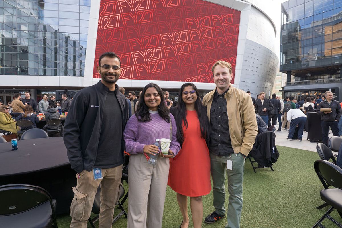 Four conference attendees pose together on outdoor terrace with modern glass buildings in background.