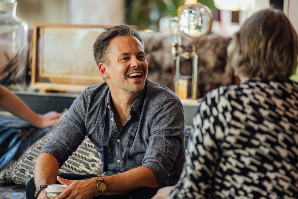 Man in gray shirt laughing while sitting on couch, talking with person in checkered pattern clothing