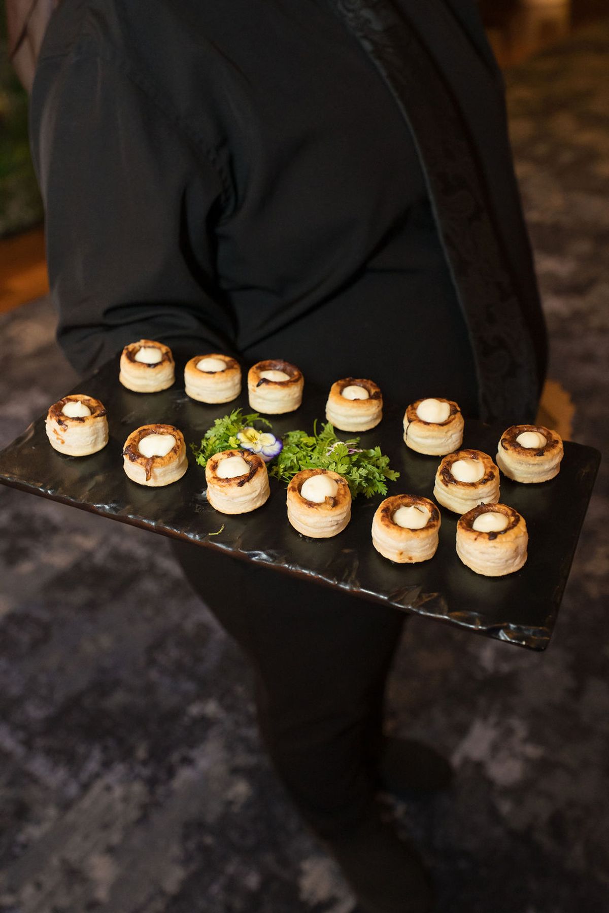 Server in black jacket holds slate tray of puff pastry appetizers topped with white cream at networking event