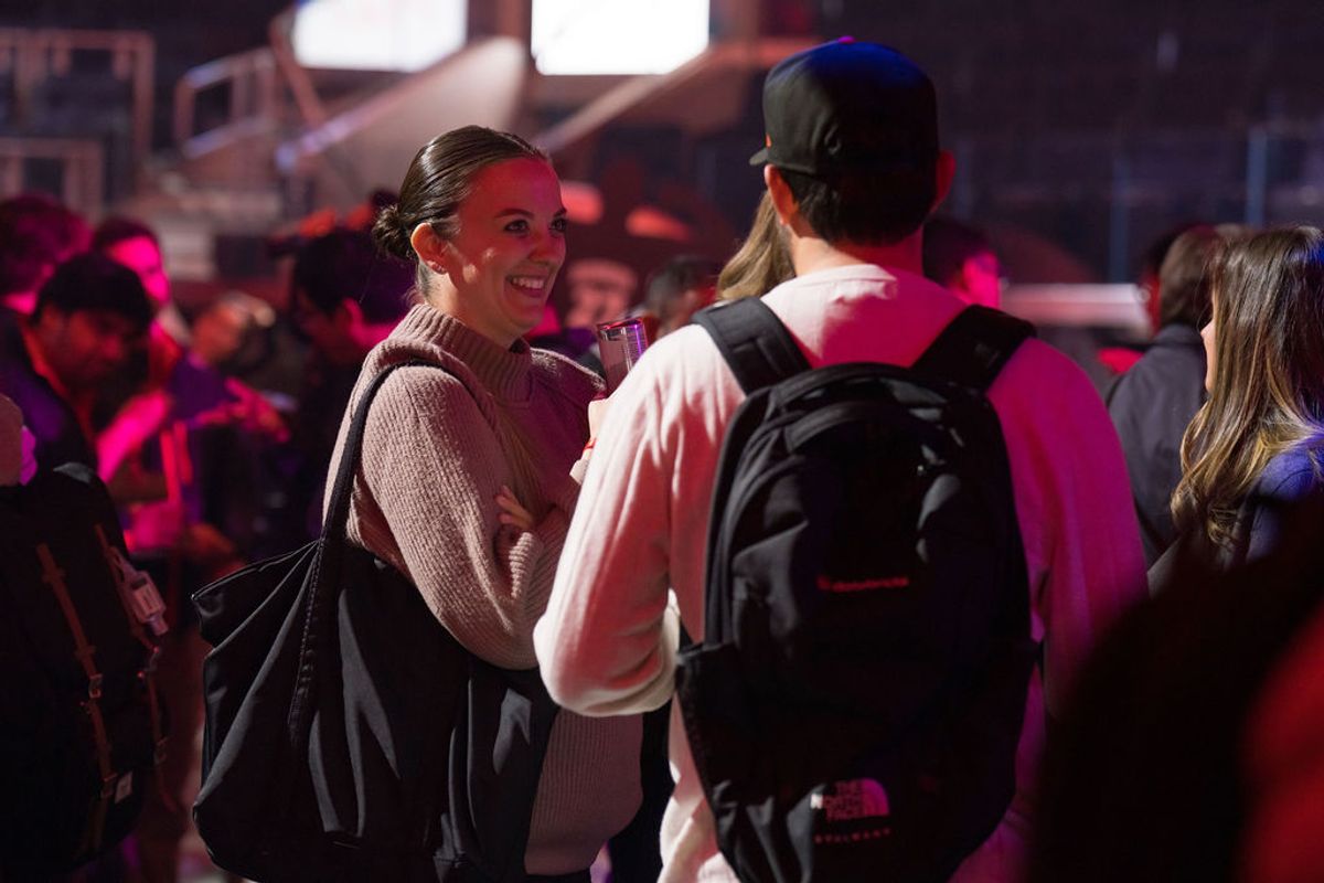 Two conference attendees smile and chat in a crowded venue with pink lighting and screens overhead