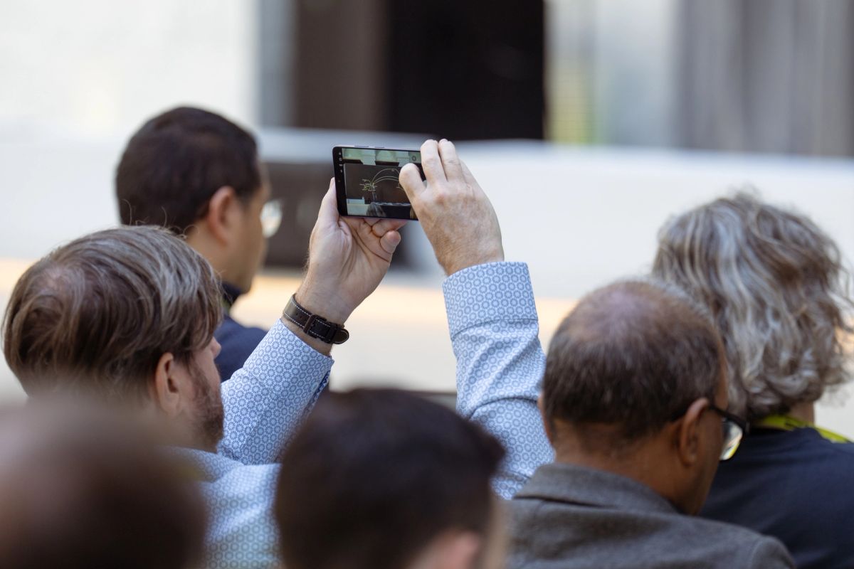 Attendee holds phone overhead to photograph a presentation among a seated crowd.
