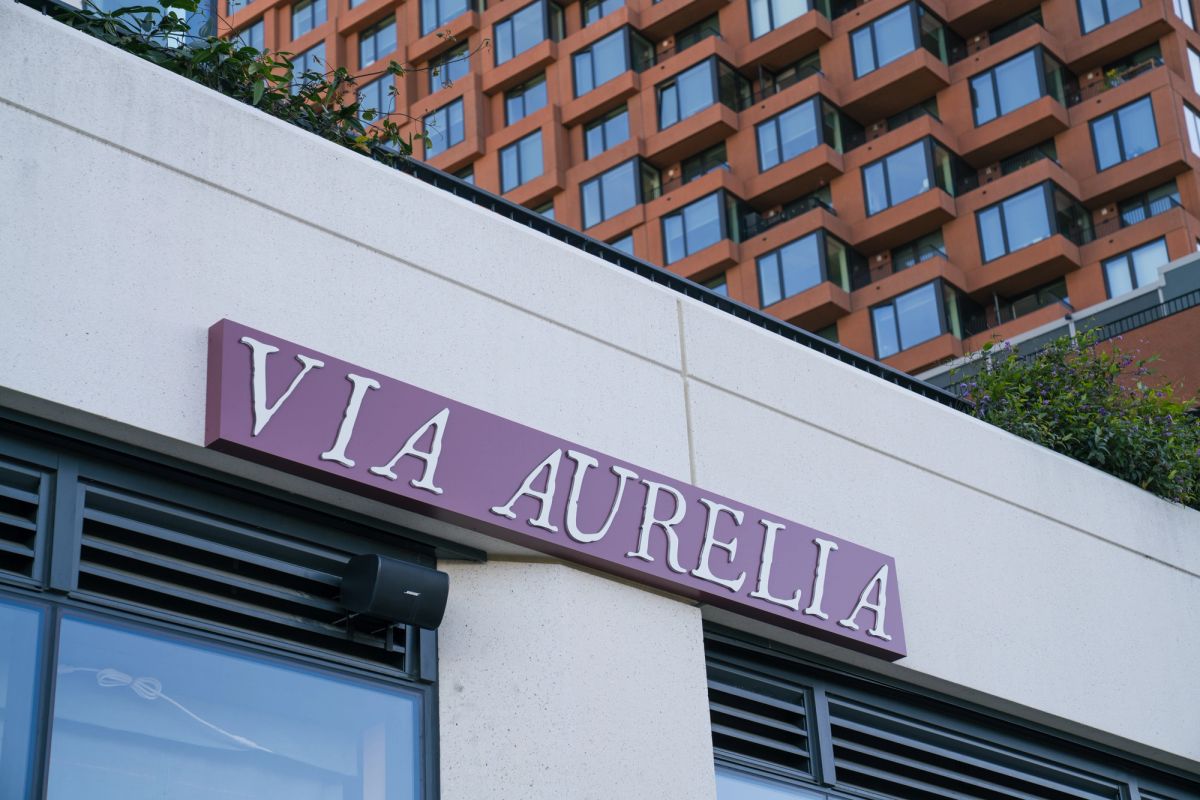 Building exterior with "VIA AURELIA" purple signage above windows, with residential tower in background
